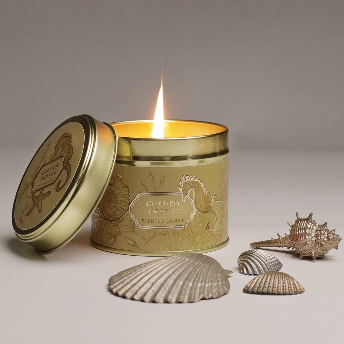 Duftkerze Golden Depths von Cream Cornwall – goldene Dose mit Seepferdchen-Illustration, brennend mit Muscheln